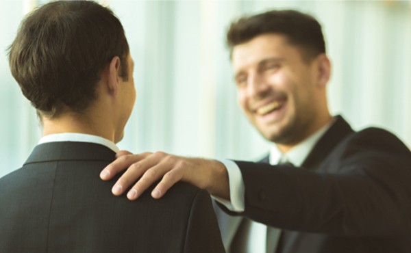 A smiling man in a suit places his hand on a second man's shoulder.