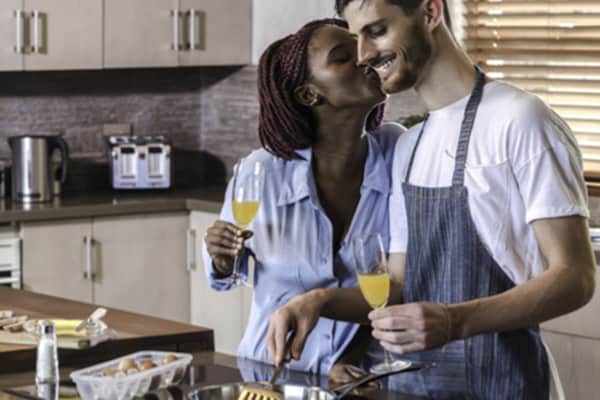 A woman holding a glass of buck's fizz kisses her partner on the cheek as he stirs a cooking pot.