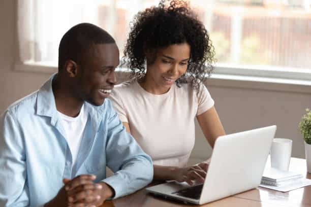 Smiling loving married mixed race couple sitting together at home with laptop, chatting with friends in messenger, planning vacation, surfing information online, shopping, watching funny videos.