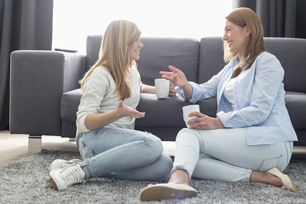 A mother and teenage daughter smile and chat as they drink coffee against a grey sofa.