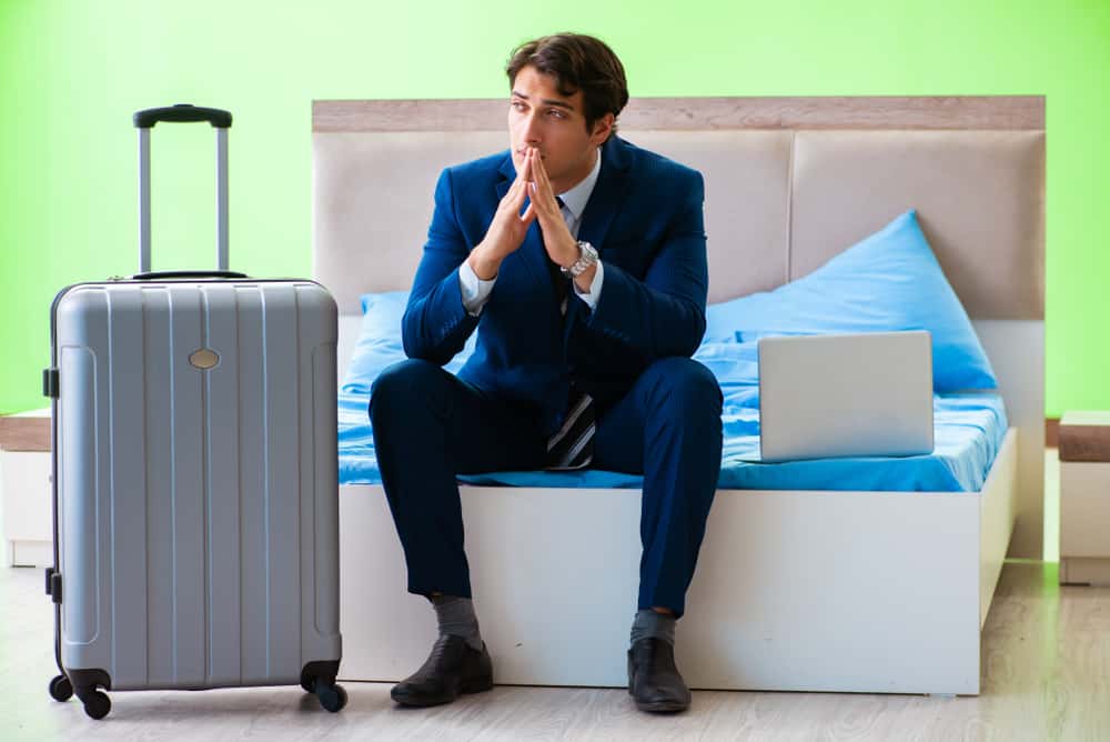 A man sits with his hands clasped under his chin at the end of a blue bed. Next to him is a wheeled grey suitcase and open white laptop.