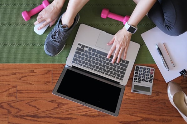 A woman in work-out gear sits on the floor surrounded by her laptop, calculator, glasses and a pen and paper.