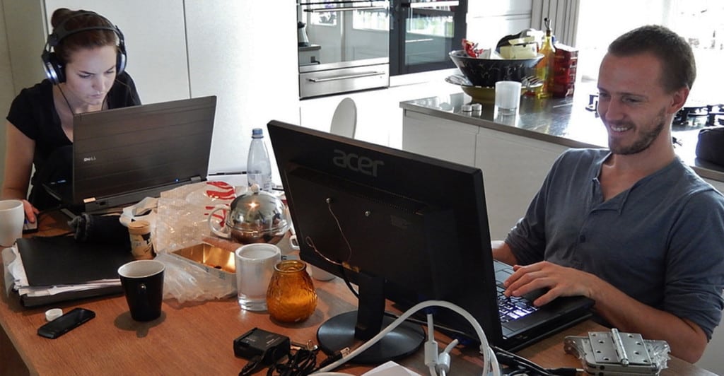 A man and woman sit around a cluttered kitchen table, each on a laptop working from home.