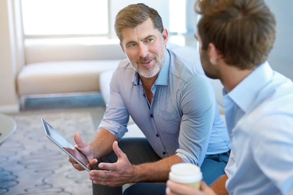 Two men in blue button up shirts sit and look at a tablet screen while drinking coffee.