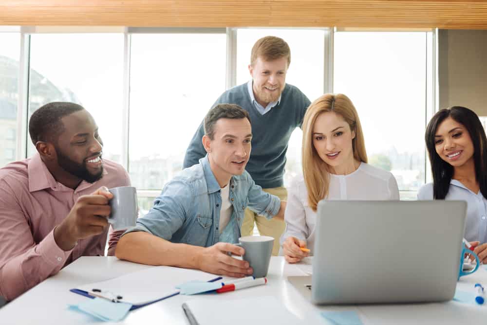 A group of 5 casually dressed office workers smile and drink coffee while looking at the same laptop screen.