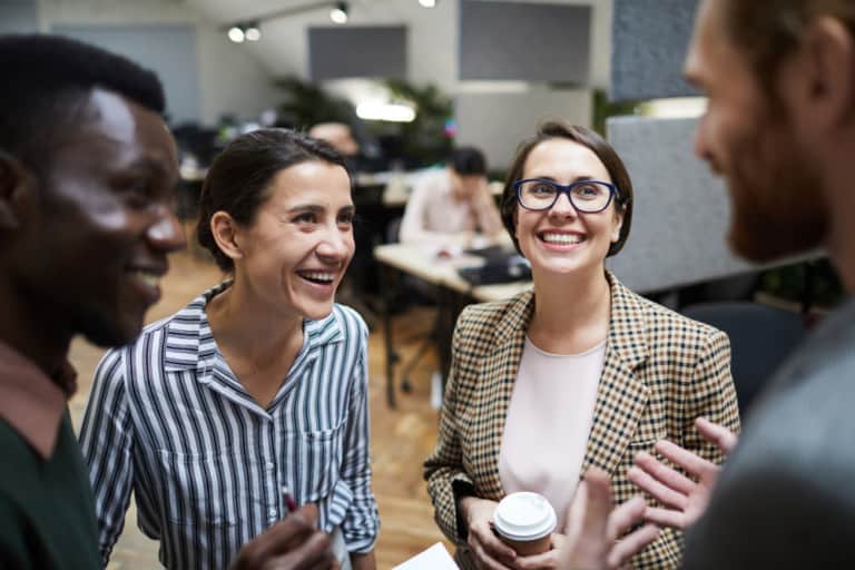 2 women both wearing smart office wear and holding coffees smile at 2 colleagues.